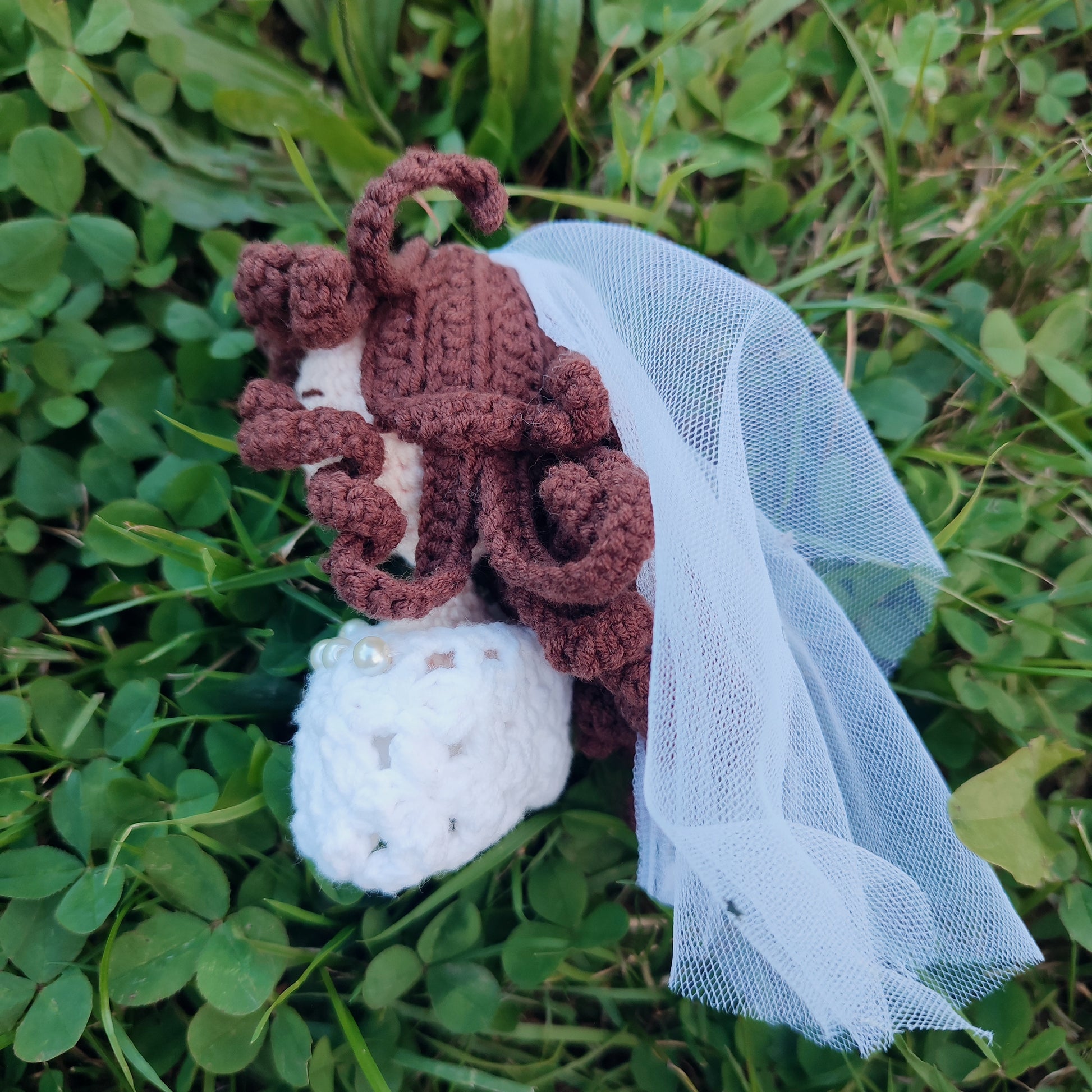 Poupée de mariée en crochet faite à la main avec des cheveux bouclés bruns, portant une robe blanche avec un collier de perles, tenue dans une main avec un fond extérieur vert.