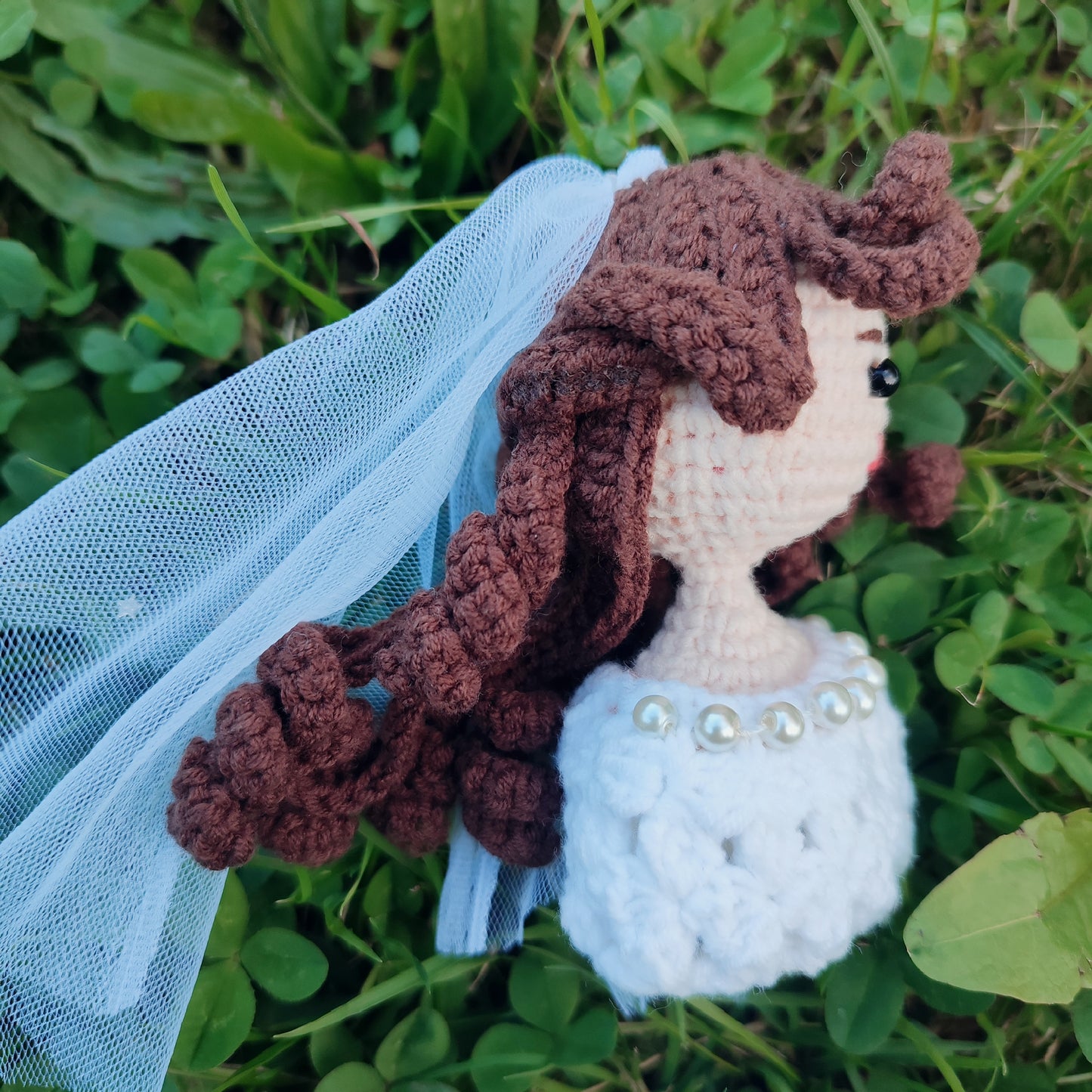 Poupée de mariée en crochet faite à la main avec des cheveux bouclés bruns, portant une robe blanche avec un collier de perles, tenue dans une main avec un fond extérieur vert.