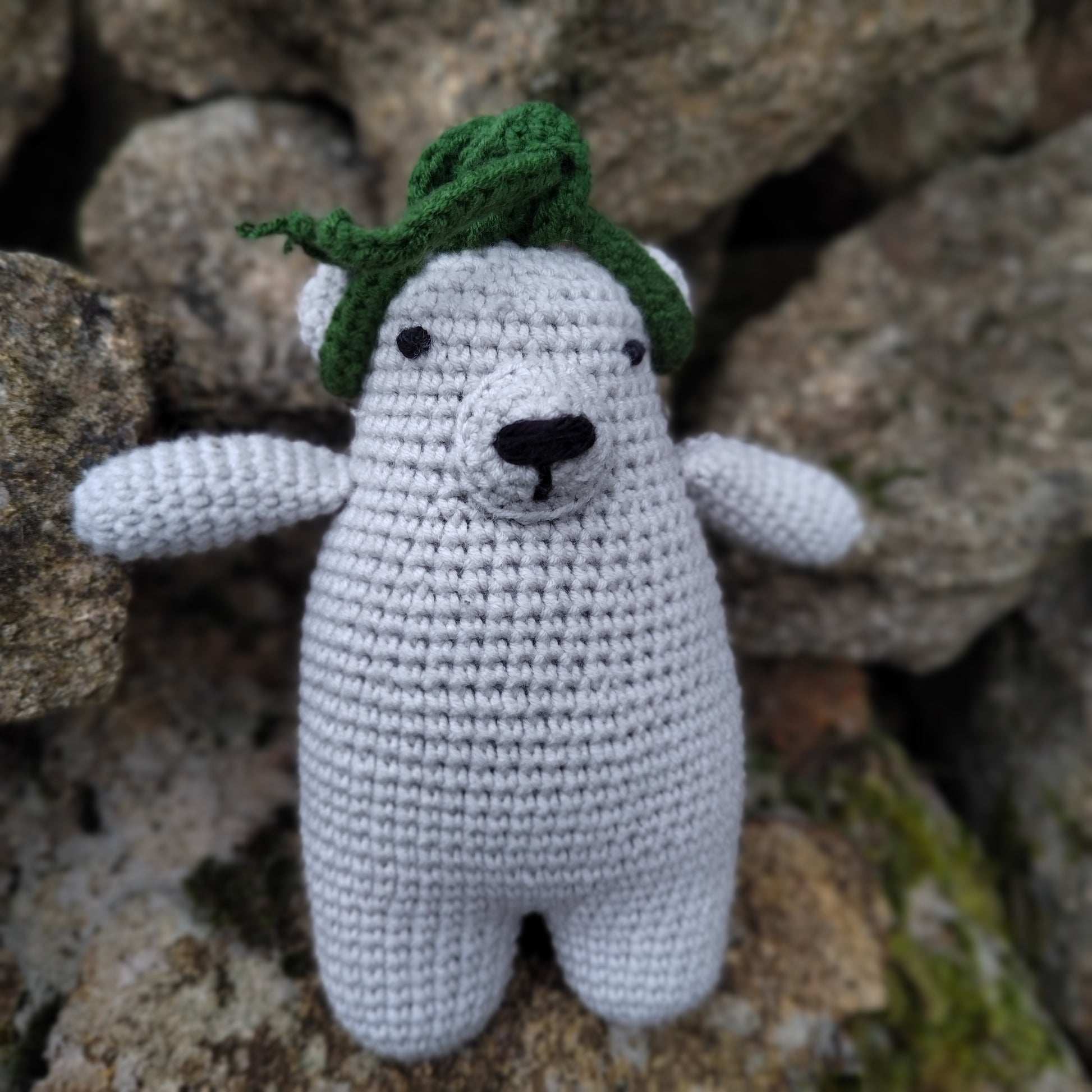 Ourson en crochet gris avec écharpe verte, posé sur des rochers naturels