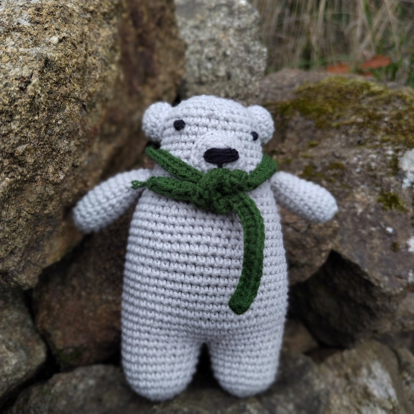 Adorable ourson en crochet gris avec une écharpe verte, debout sur un fond de rochers.Adorable ourson en crochet gris avec une écharpe verte, debout sur un fond de rochers.