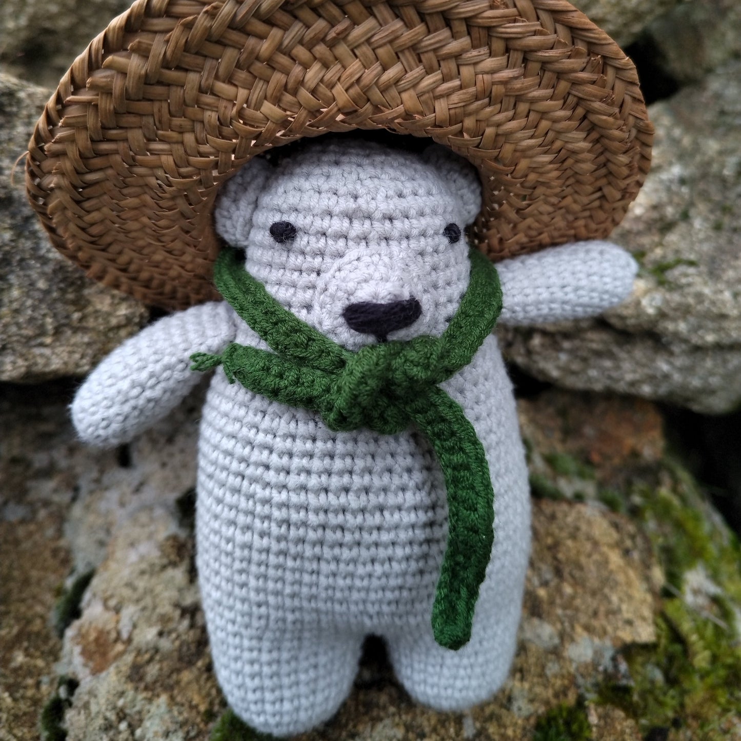 Ours en crochet gris aux détails soignés, portant une écharpe verte, photographié sur des rochers