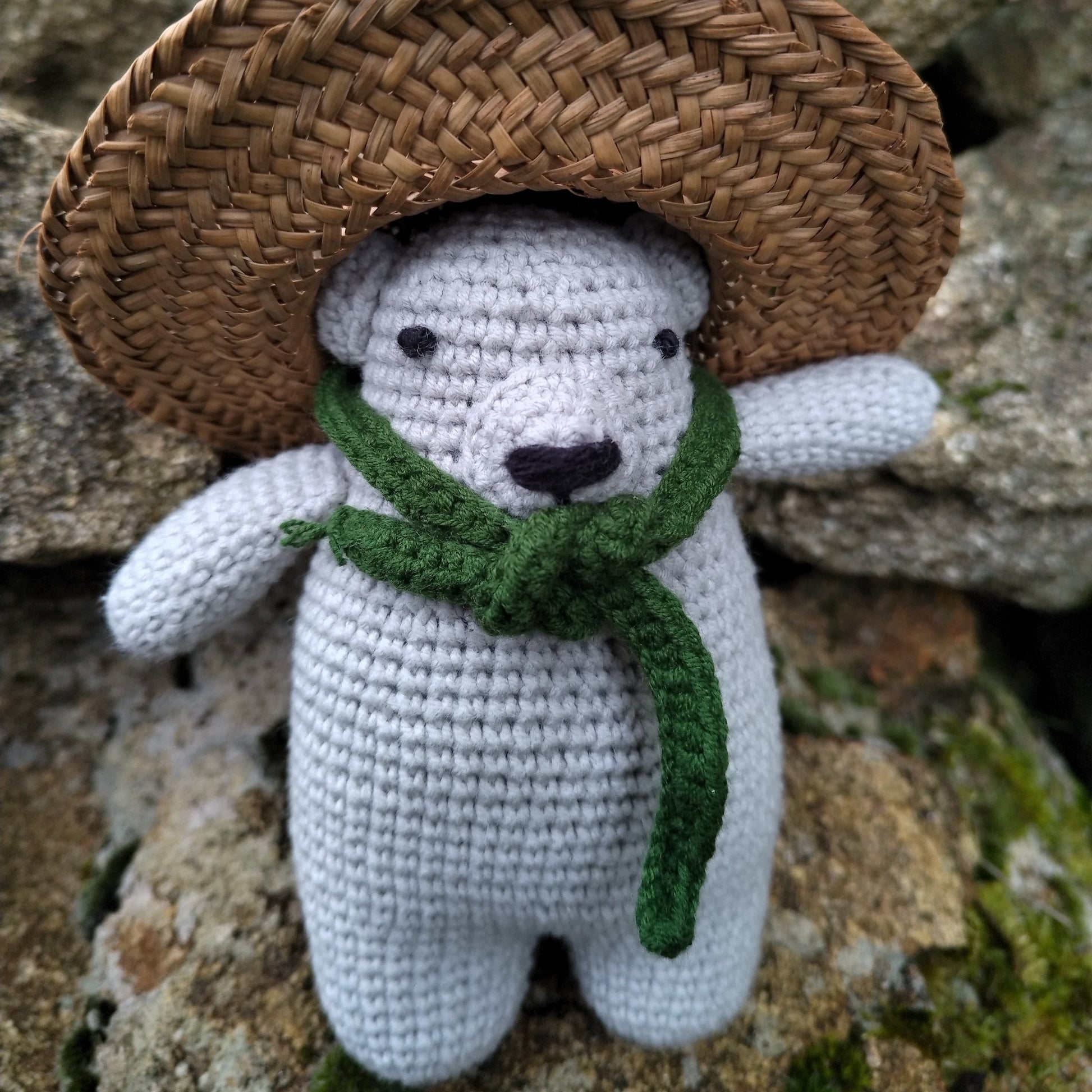 Ours en crochet gris aux détails soignés, portant une écharpe verte, photographié sur des rochers