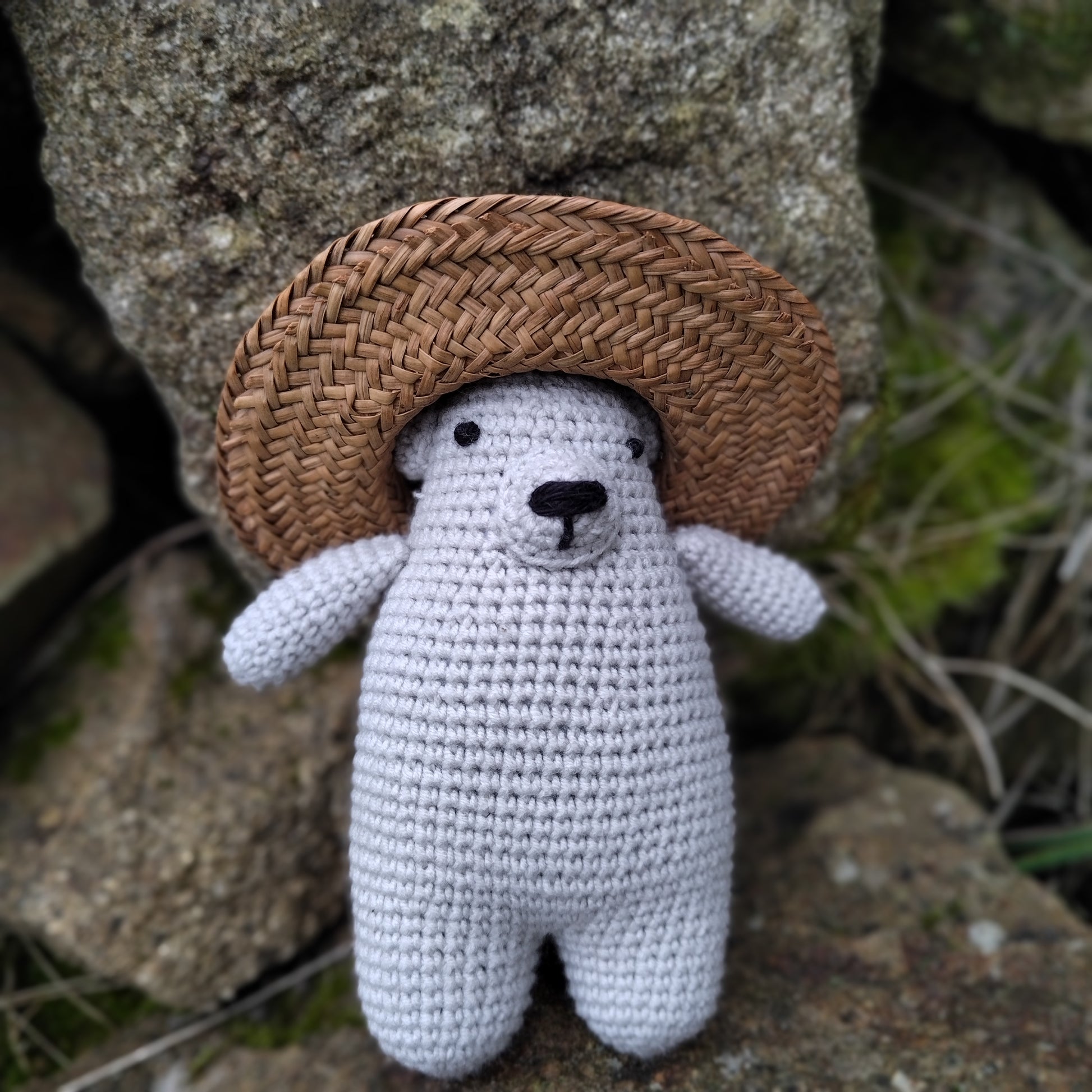 Petit ourson en crochet gris avec écharpe verte, posé sur des rochers, prêt à être adopté